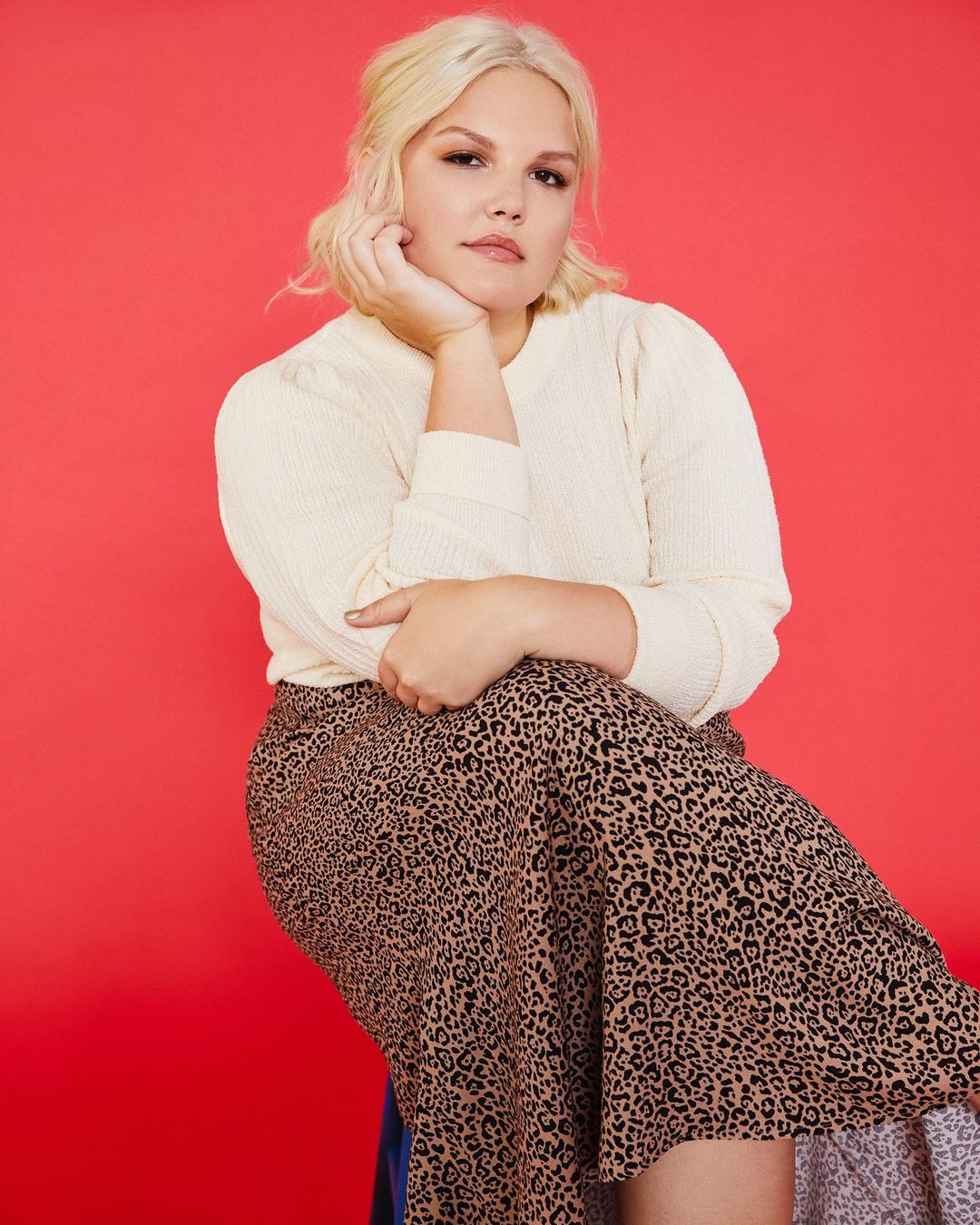 Woman in cream sweater and leopard print skirt poses in front of red background in a beauty or fashion shot. 