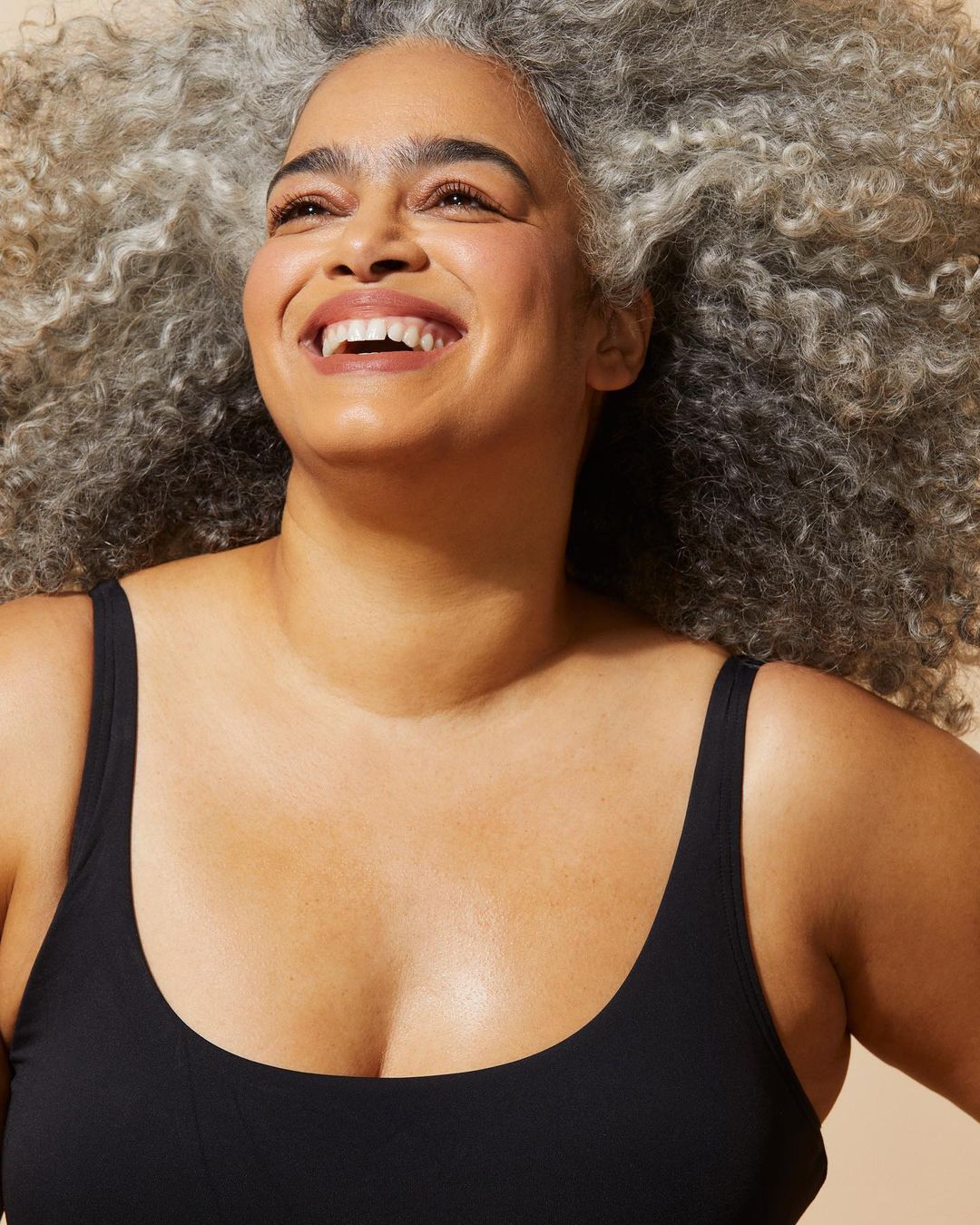 Smiling adult woman with curly gray hair in a black top against a tan background, portrait-style studio shot, beauty and skincare concept.