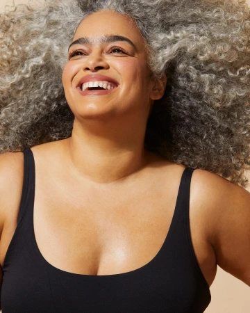 Close-up portrait of a smiling woman with voluminous curly grey hair wearing a black tank top against a plain background