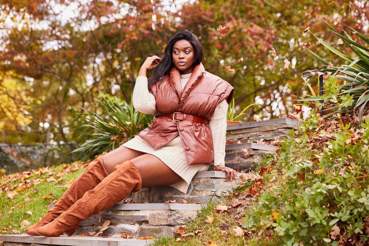 Autumn fashion portrait of a woman in a puffer vest and thigh-high boots sitting on stone steps in a park surrounded by fall foliage.