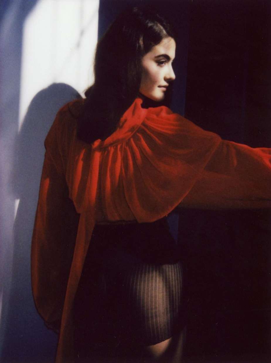 Indoor fashion shot of a woman in a red top and dark skirt posing near a sunlit wall with shadow, showcasing studio lighting.