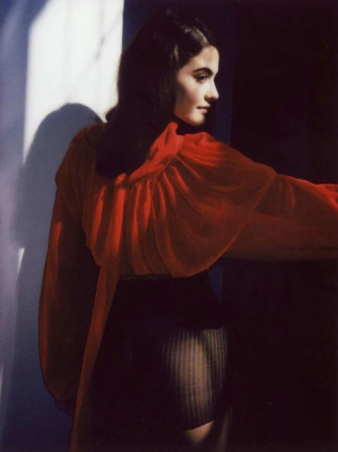Indoor fashion shot of a woman in a red top and dark skirt posing near a sunlit wall with shadow, showcasing studio lighting.