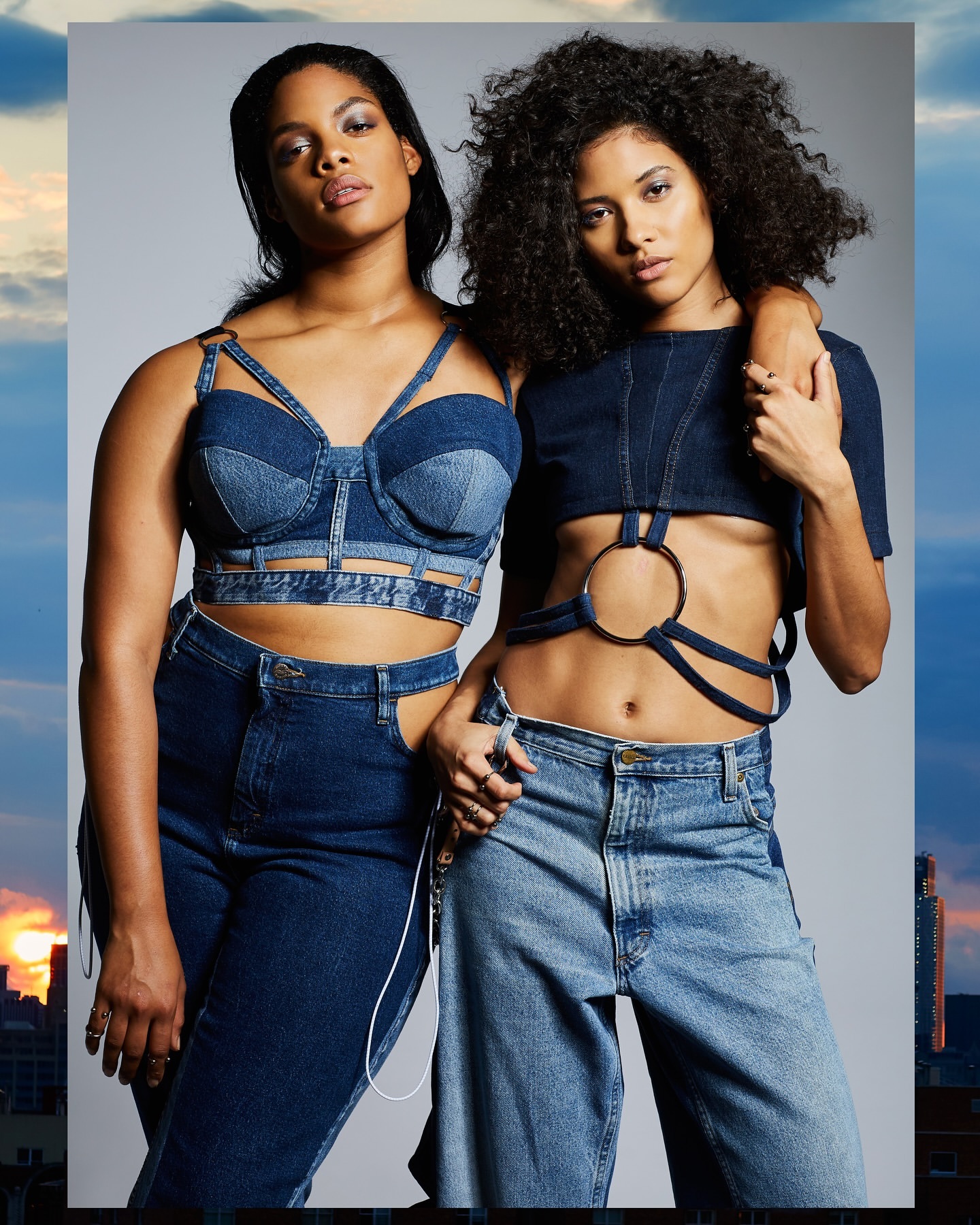 Two adult women pose in matching denim outfits, showcasing a modern fashion trend with natural hair against a grey backdrop.