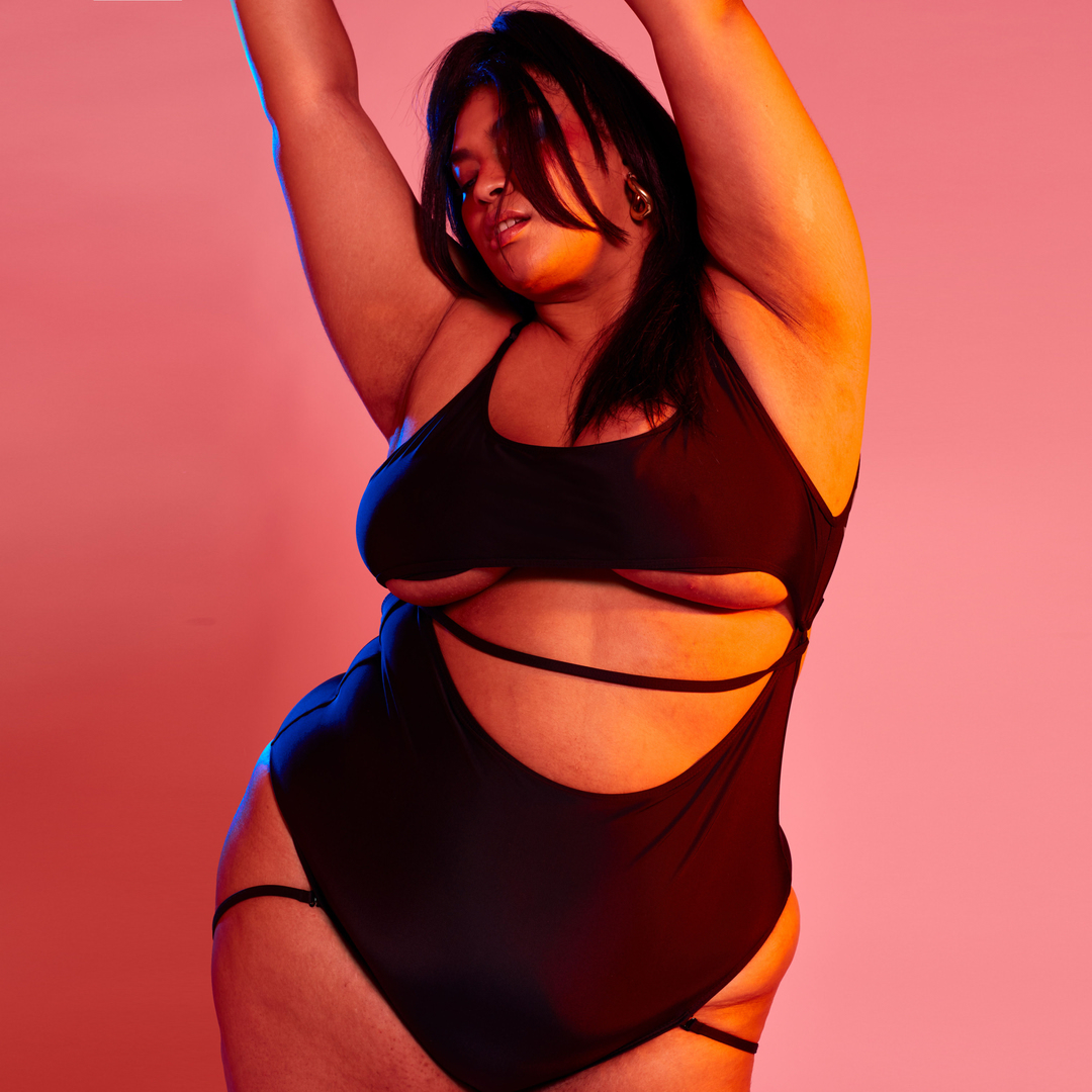 Adult woman in black bodysuit strikes a sensual pose in a pink photo studio illuminated by colored lights, creating a visually arresting image.