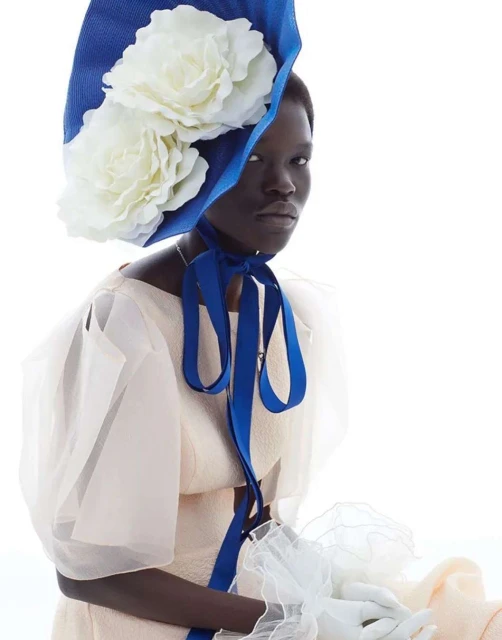 Fashionable portrait of an adult woman wearing a large, elegant hat with white floral accents, with a pure white background.