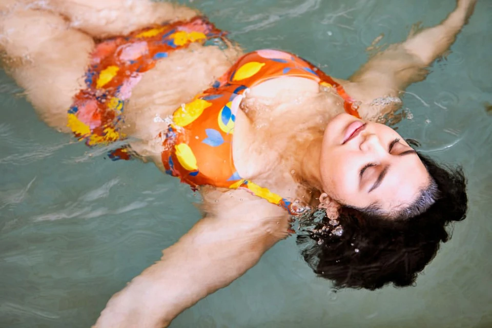 Woman floats in water wearing a patterned bikini. Close-up shows her face, arms, and chest with water surrounding her. Outdoor scene, eye-level view.