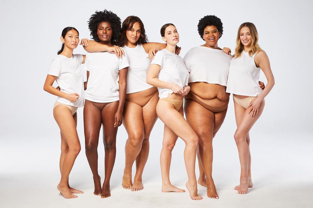 Six diverse women pose in a studio in white tops and neutral bottoms, showcasing a body-positive fashion campaign with a bright, clean aesthetic.