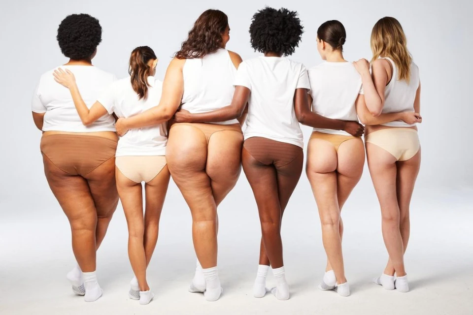 Diverse group of six women wearing white shirts and neutral-colored underwear, standing close together with arms around each other's shoulders.