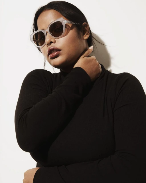 Close-up studio shot of a plus-size woman with fair skin wearing sunglasses, a black long-sleeve shirt, and holding her neck with her hand.