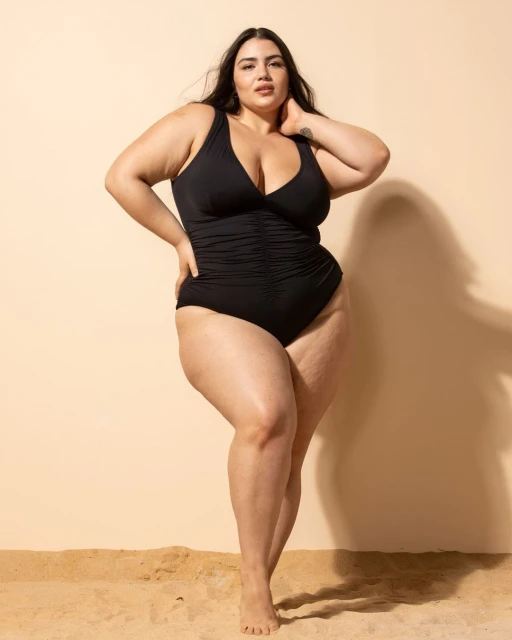 Plus-size woman in a black one-piece swimsuit posing confidently against a neutral backdrop with a sandy surface, showcasing body positivity.