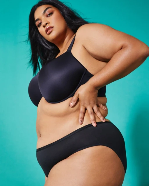 Side view of an adult female model in black bra and underwear posing indoors against a teal background, showing her mid-section.