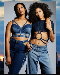 Two women posing together wearing matching blue denim crop tops and high-waisted jeans against a grey studio backdrop.