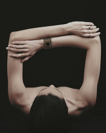Close-up portrait of a person with arms crossed forming a frame around the head, wearing a wide gold watch and rings against a dark background.