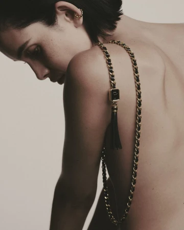 Close-up portrait of a woman wearing a gold chain strap watch with black leather detail draped over her bare shoulder.