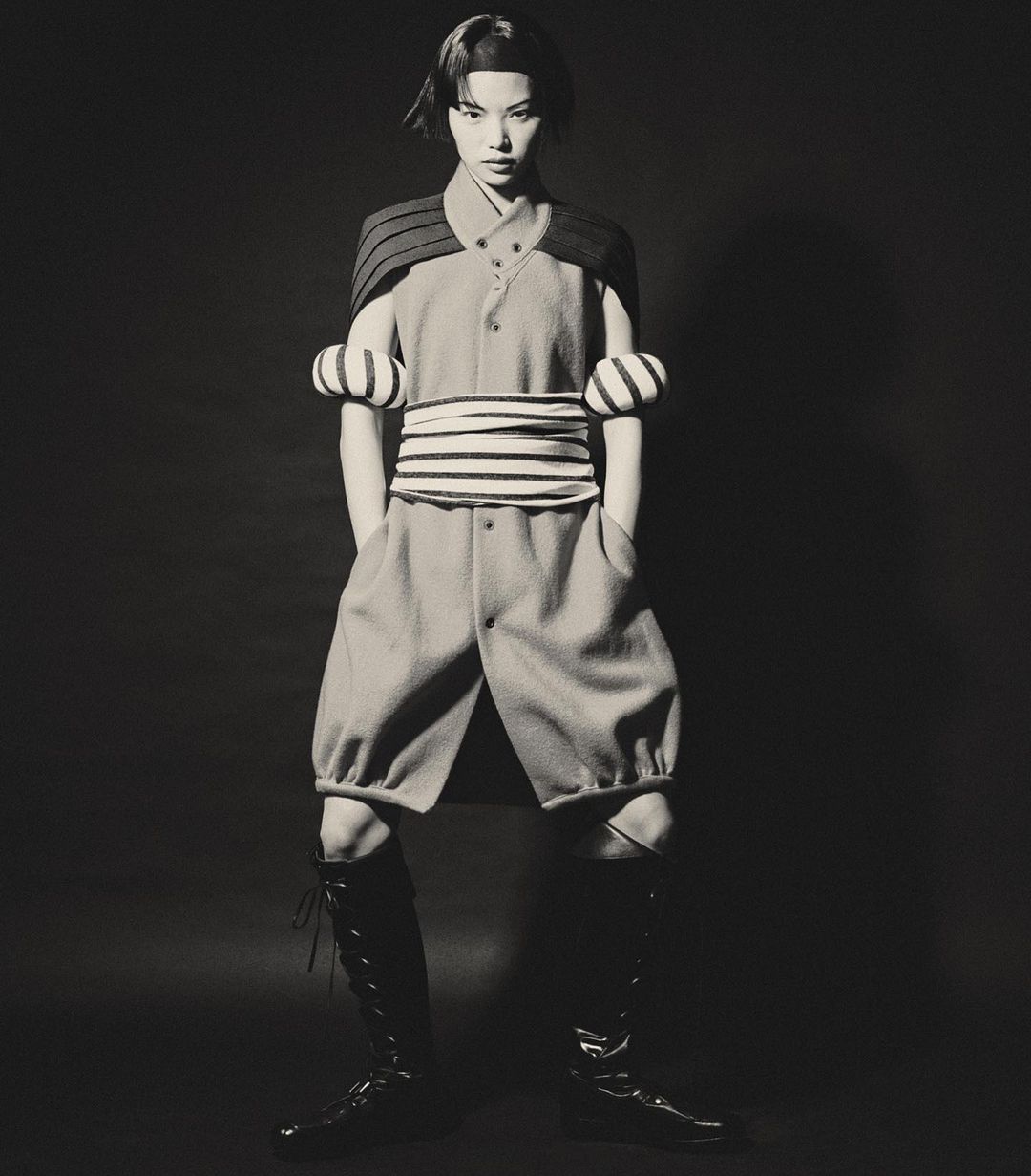A person in a light blue outfit with black trim and high boots stands in a studio with a dark background in a fashion shot.