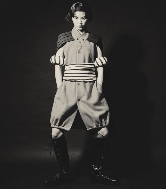 A person in a light blue outfit with black trim and high boots stands in a studio with a dark background in a fashion shot.