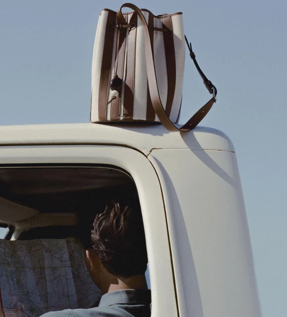 Striped canvas and leather bucket bag on top of a vintage camper van under a clear sky, with a man looking at a map inside.