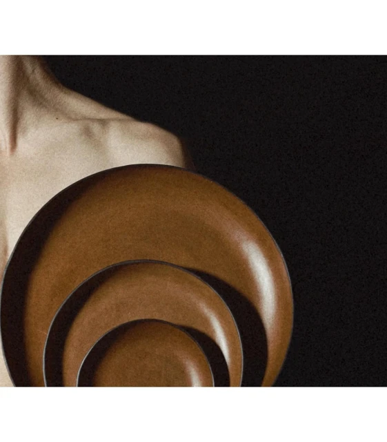 Three brown ceramic plates are stacked in descending size next to the bare shoulder of an adult with a dark background.