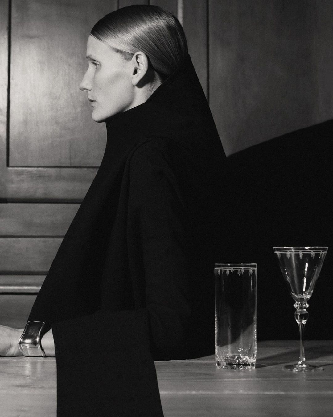 Elegant profile of woman in black turtleneck with minimalist jewelry. Two glasses are placed on a table with a dark background.