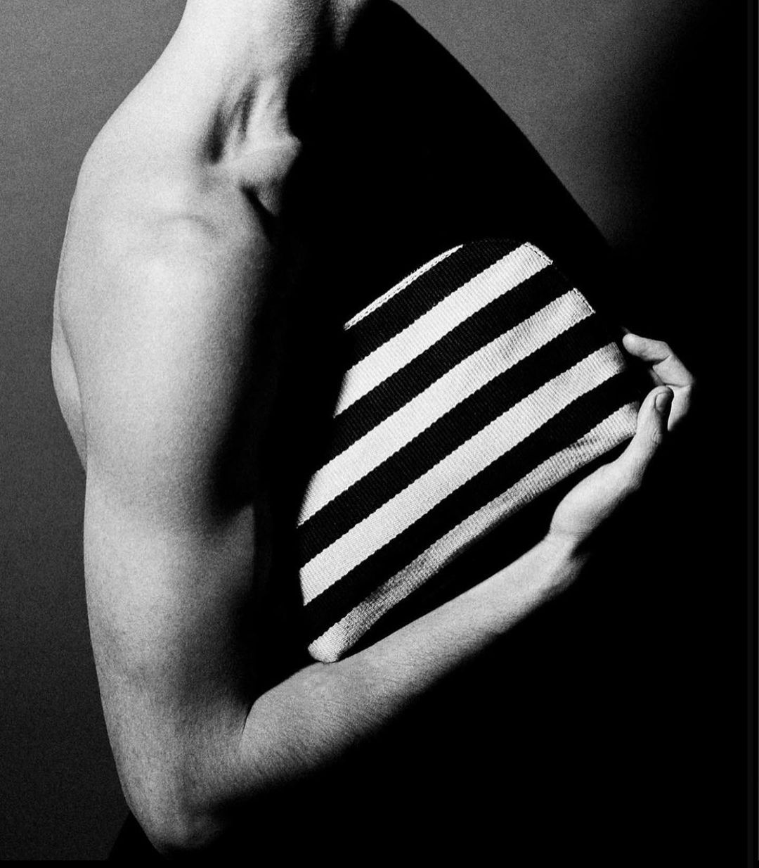 Black and white studio shot of a fair-skinned adult holding a striped cushion, showcasing minimalist aesthetics and body form.