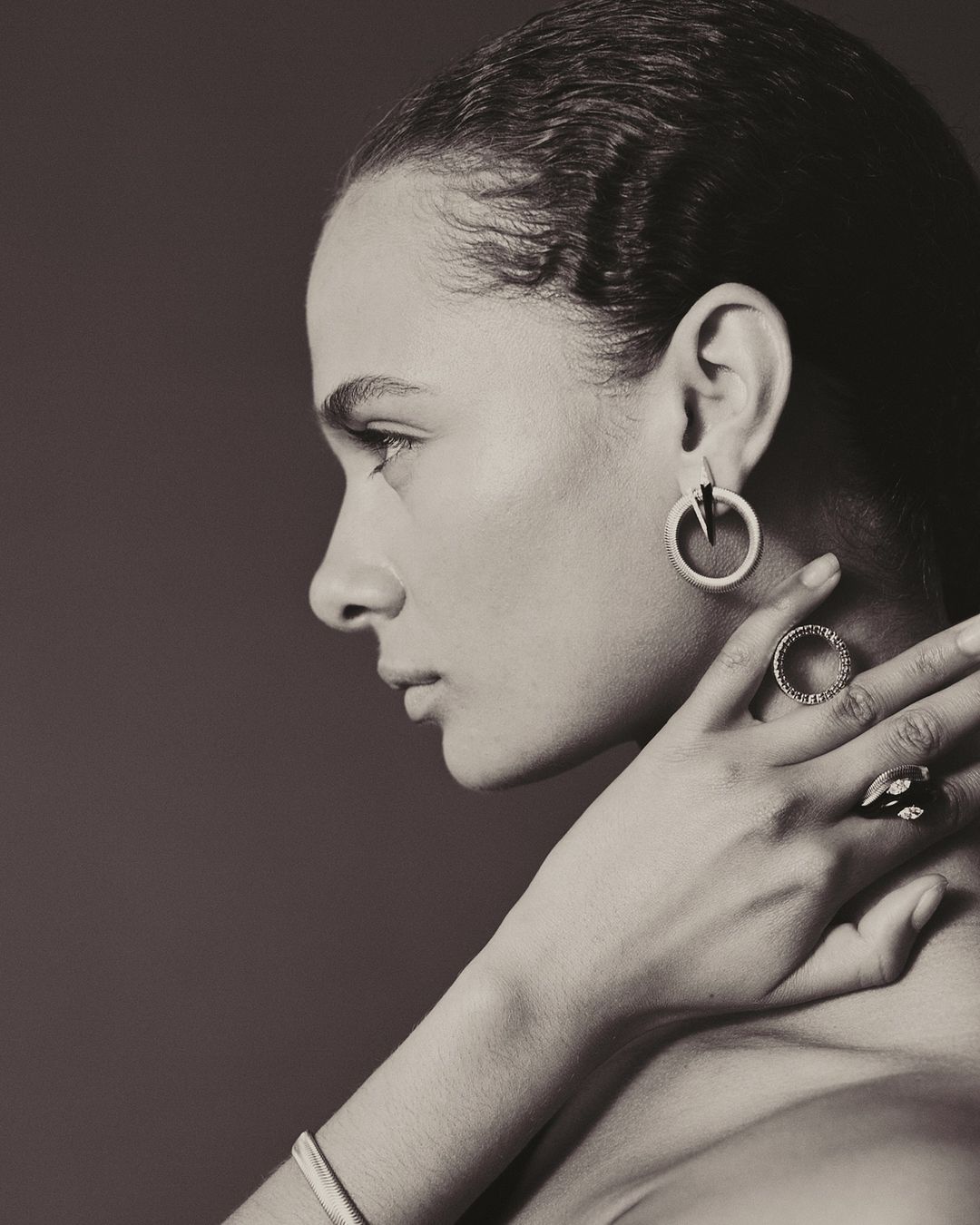 Profile portrait of a woman in monochrome, wearing sculptural hoop earrings and rings while touching her neck.