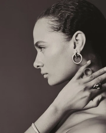 Profile portrait of a woman in monochrome, wearing sculptural hoop earrings and rings while touching her neck.