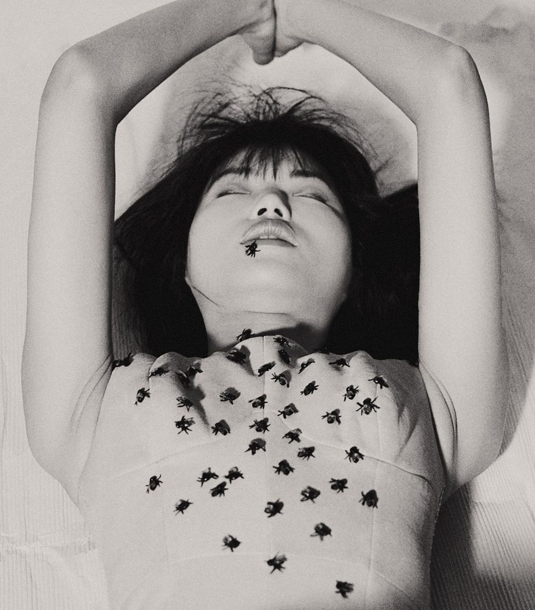 Black and white portrait of a woman lying on a white surface, adorned with fly-shaped decorations on her top and a fly on her lips.