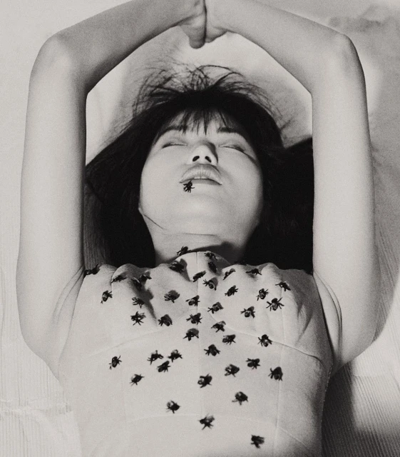 Black and white portrait of a woman lying on a white surface, adorned with fly-shaped decorations on her top and a fly on her lips.