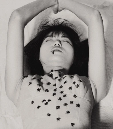 Black and white portrait of a woman lying on a white surface, adorned with fly-shaped decorations on her top and a fly on her lips.