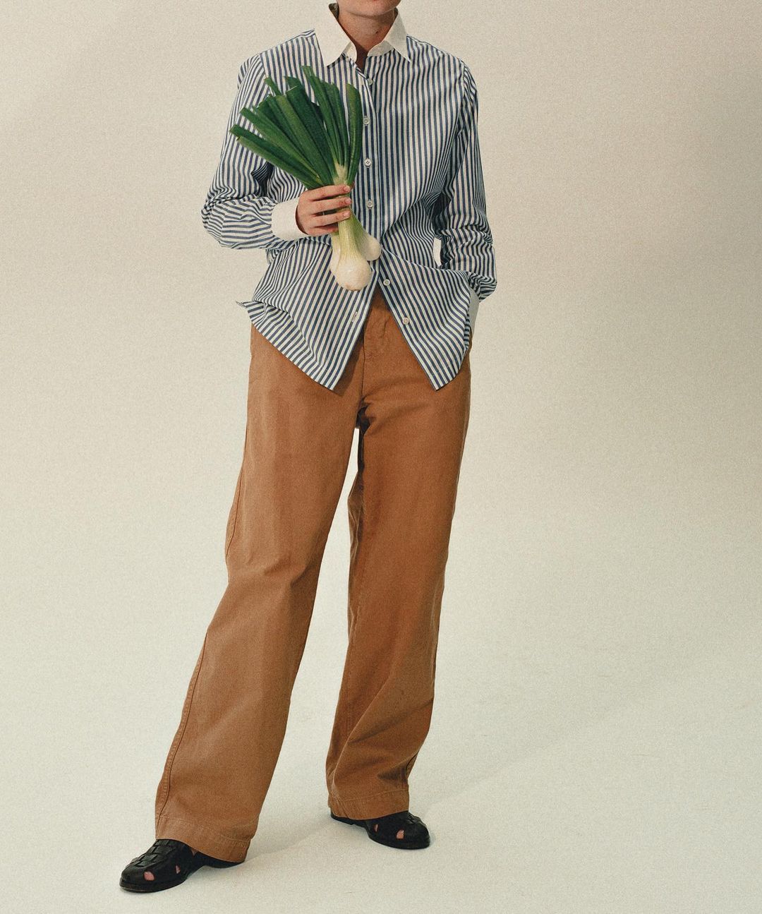 A stylish person in a striped shirt and tan pants holds fresh green onions in a studio setting with soft, neutral lighting.