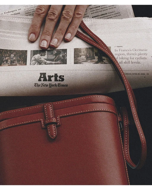 Close-up of a red leather shoulder bag and a hand resting on a newspaper featuring articles and images from The New York Times Arts section.