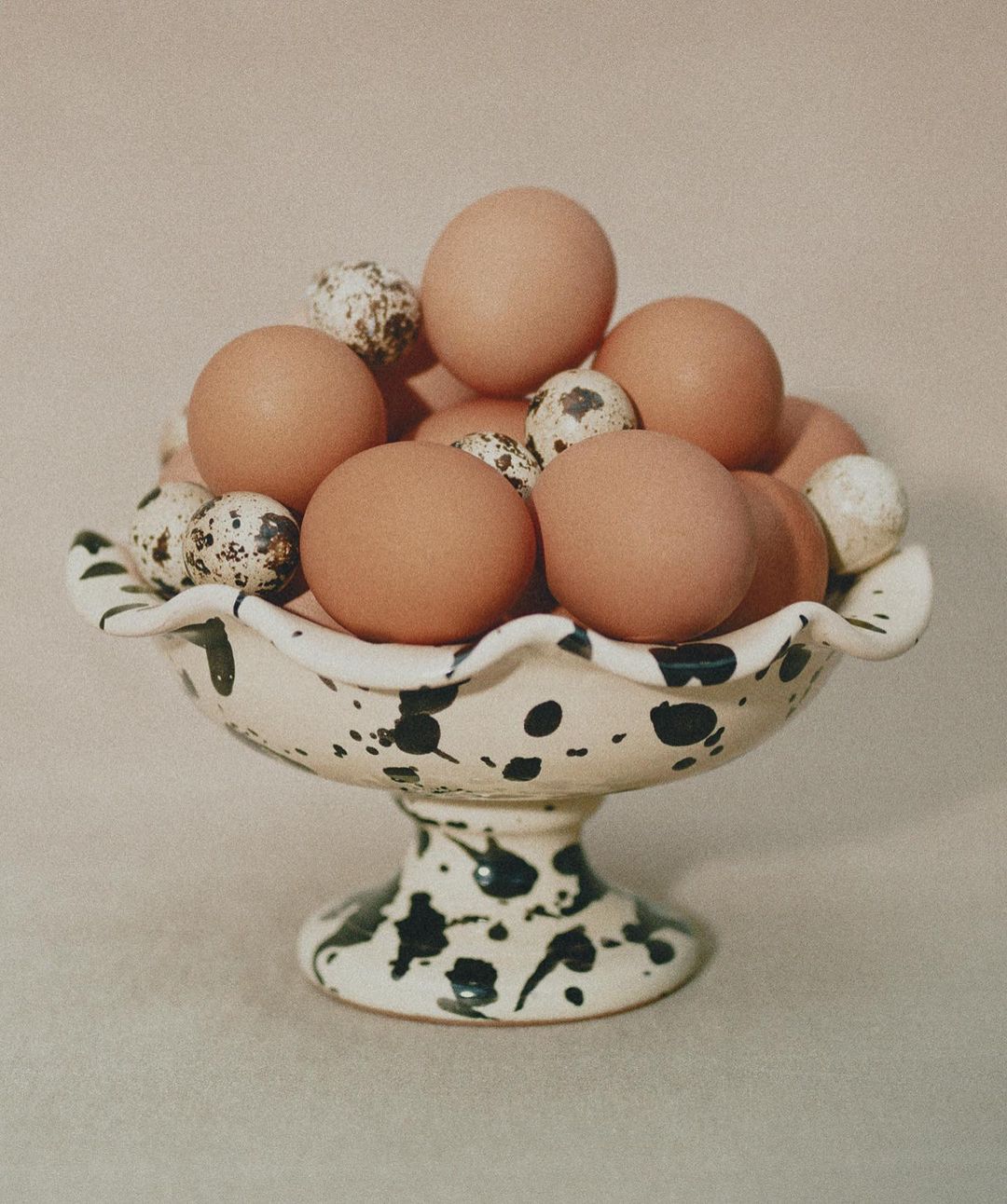 Still life of brown chicken eggs and speckled quail eggs in a decorative white and black speckled pedestal bowl on a neutral surface.