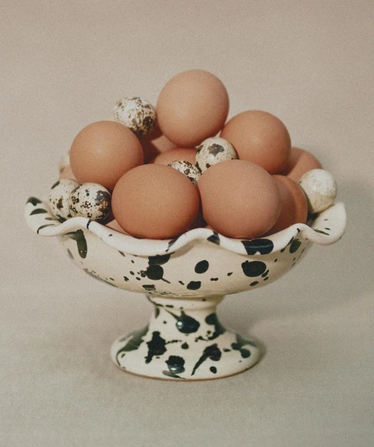 Still life of brown chicken eggs and speckled quail eggs in a decorative white and black speckled pedestal bowl on a neutral surface.