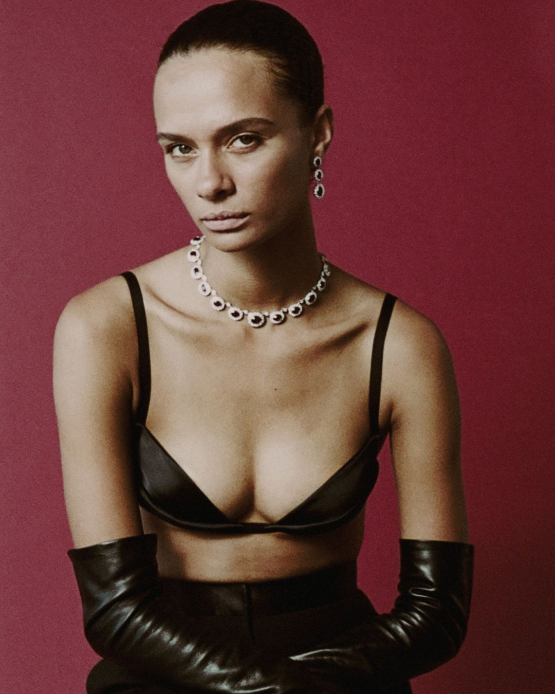 A woman poses in black bra, pants and gloves with jewelry against a maroon background, showcasing a minimalist fashion portrait.