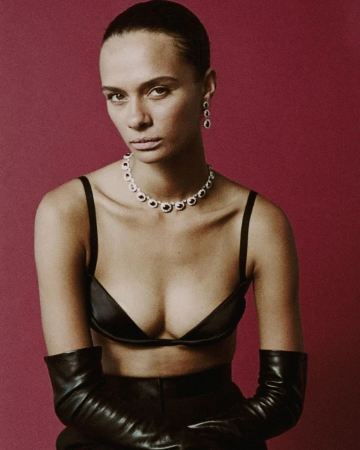 A woman poses in black bra, pants and gloves with jewelry against a maroon background, showcasing a minimalist fashion portrait.
