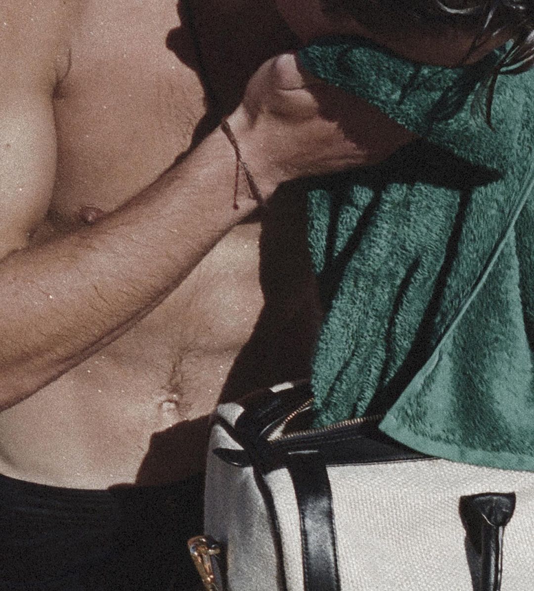 Sweaty man's torso with abs, wearing a string bracelet, drying with a green towel, and a white duffel bag in a close-up shot.