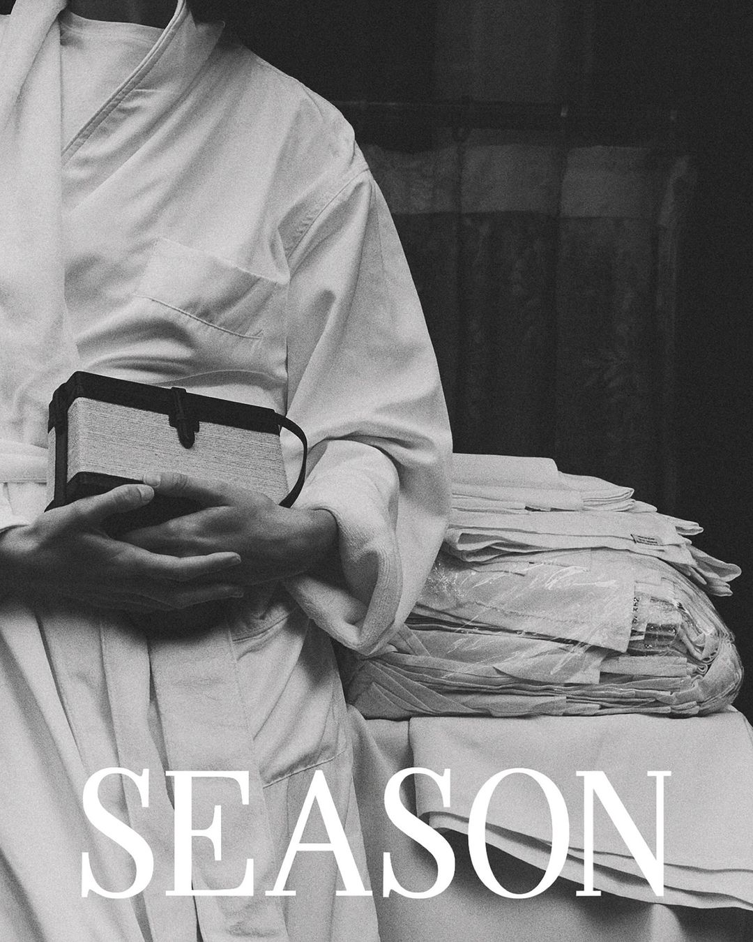 Monochrome fashion portrait by Anastasiia Duvallie showing a person in a white robe holding a structured handbag.