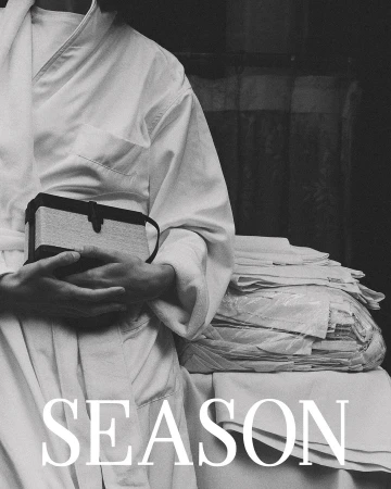 Monochrome fashion portrait by Anastasiia Duvallie showing a person in a white robe holding a structured handbag.