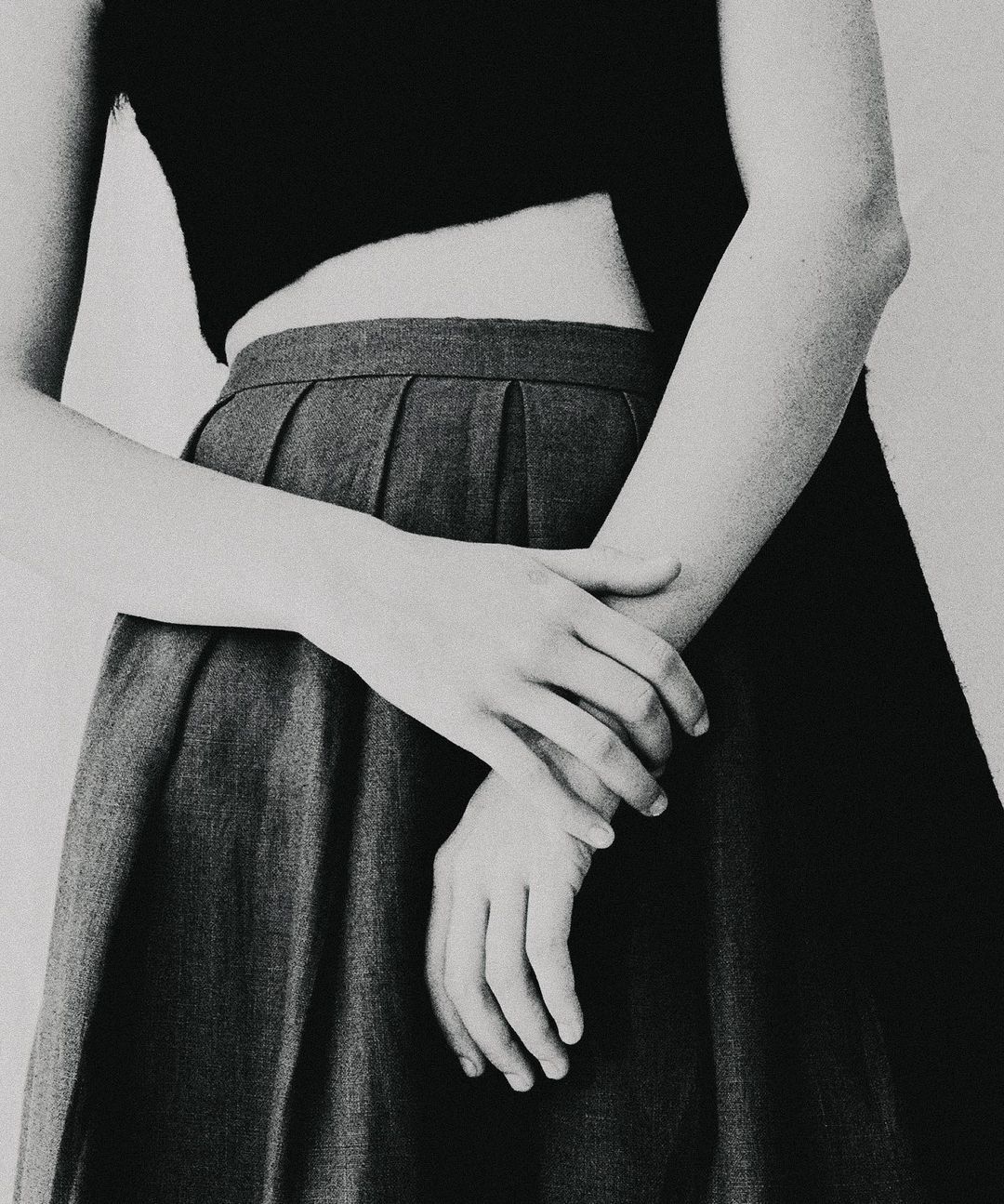 Monochrome close-up of a young adult with crossed arms, wearing a black crop top and gray pleated skirt against a white background.