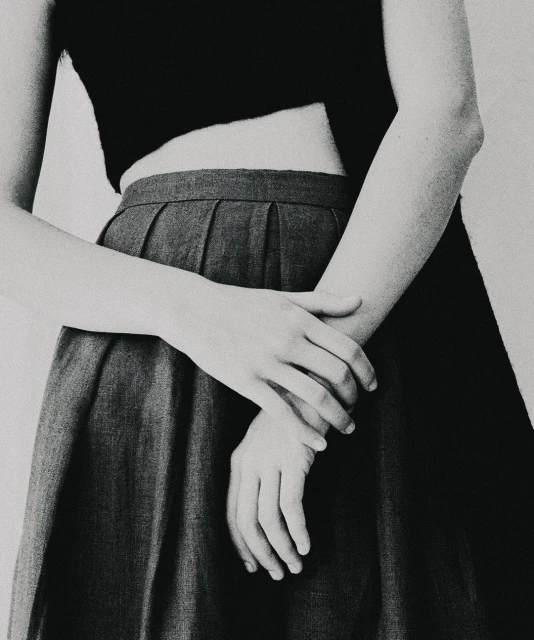 Monochrome close-up of a young adult with crossed arms, wearing a black crop top and gray pleated skirt against a white background.