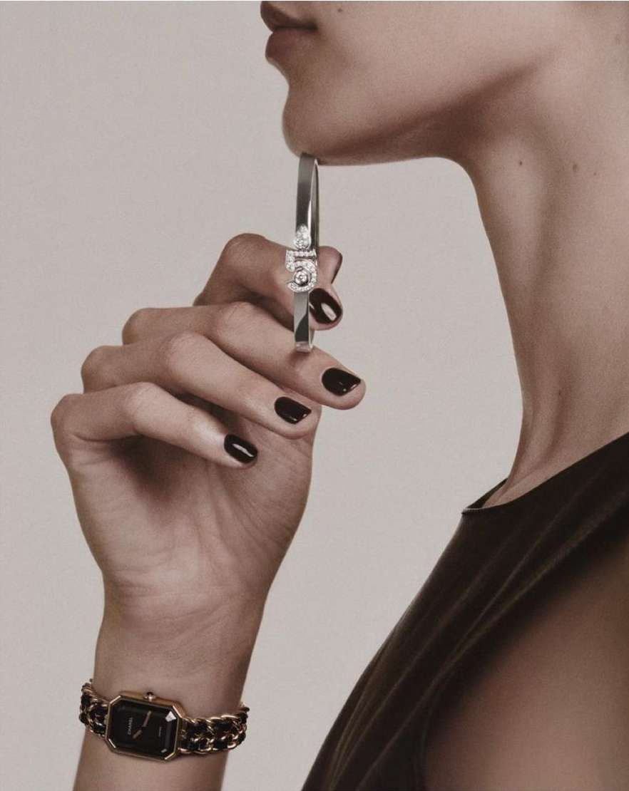 Closeup of a woman wearing luxury jewelry, including a silver bracelet and a black watch with a gold chain, against a neutral background.