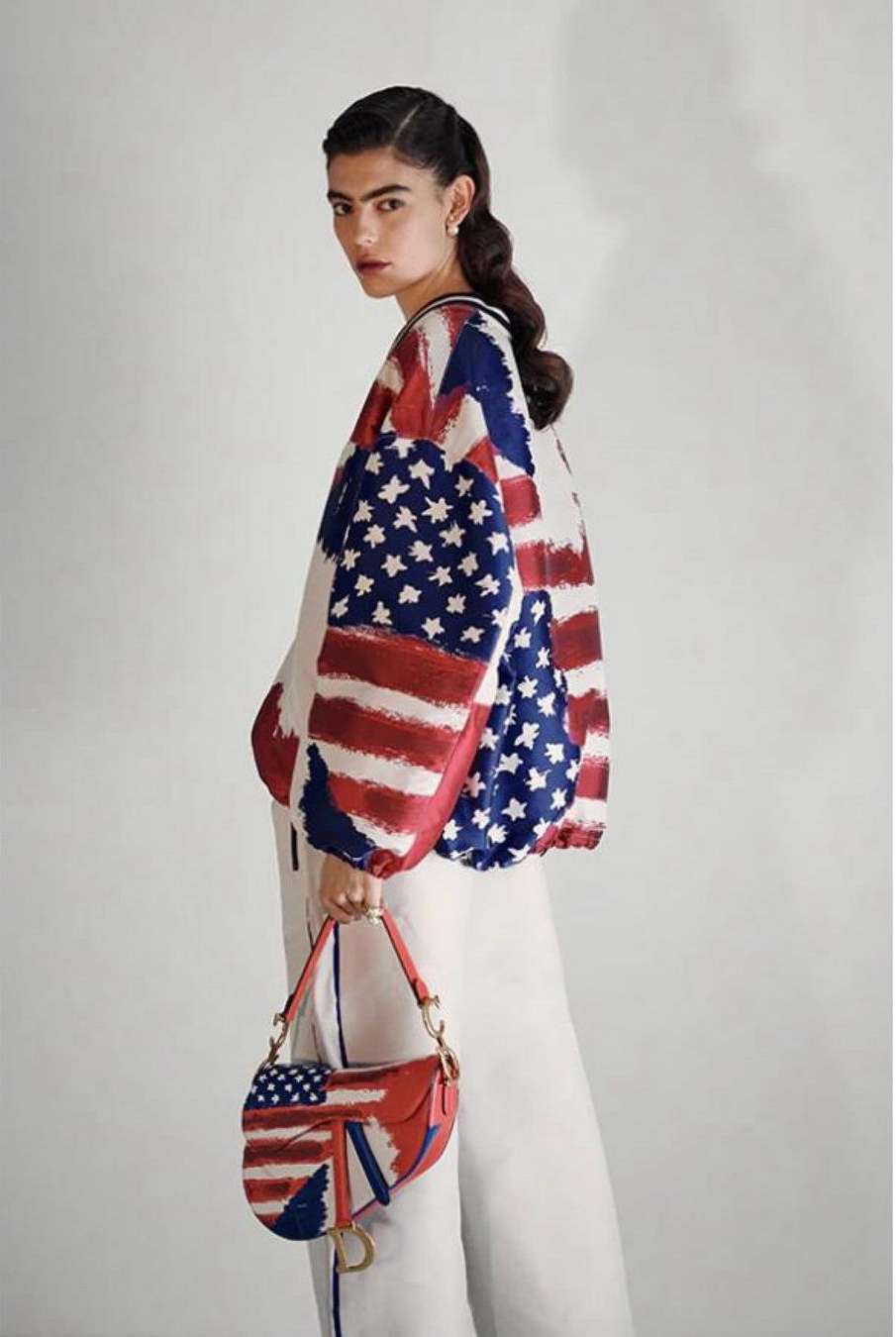Woman in American flag-themed jacket and white pants holding a matching handbag against a plain studio backdrop for fashion editorial.