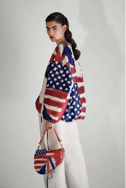 Woman in American flag-themed jacket and white pants holding a matching handbag against a plain studio backdrop for fashion editorial.
