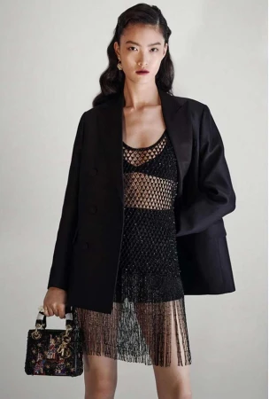 Model in black blazer and embellished mesh dress holding a small dark handbag against a plain background