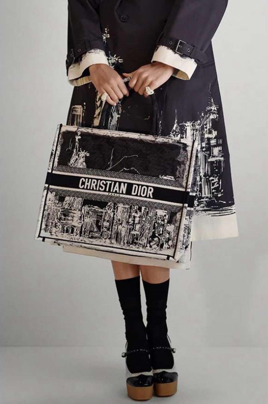 Fashion shot: Woman with Christian Dior tote bag, wearing black and white coat, knee socks, and platform heels in studio.