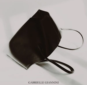 A dark structured leather tote bag rests at an angle against a bright white seamless background with thin black straps.