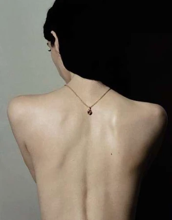 Rear view of a person with dark hair wearing a delicate gold pendant necklace against a split-tone light and dark background.