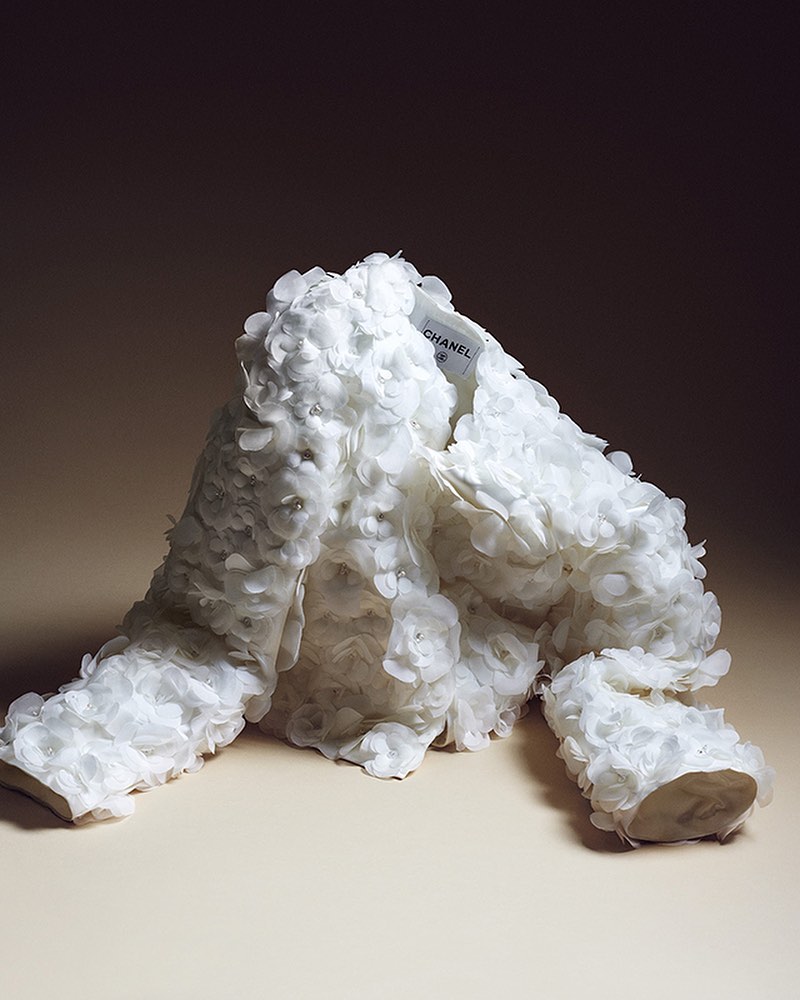 A white, flower-covered Chanel jacket on a beige background, showcasing high fashion and textile artistry in a studio setting.