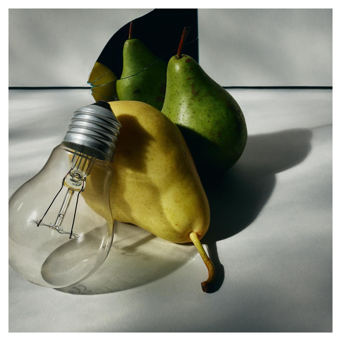Still life photograph of a lightbulb and two pears, one green and one yellow, set against a white surface with a dark shadow.