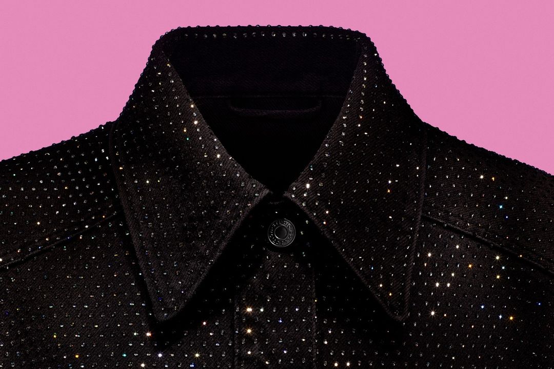 Close-up of a black denim jacket collar, adorned with sparkling rhinestones, set against a light pink background for fashion appeal.